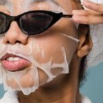 Routines - Self esteem young African American lady with moisturizing sheet mask and towel on hand wearing trendy sunglasses while standing against blue background in bathrobe after shower