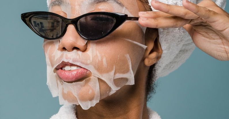 Routines - Self esteem young African American lady with moisturizing sheet mask and towel on hand wearing trendy sunglasses while standing against blue background in bathrobe after shower